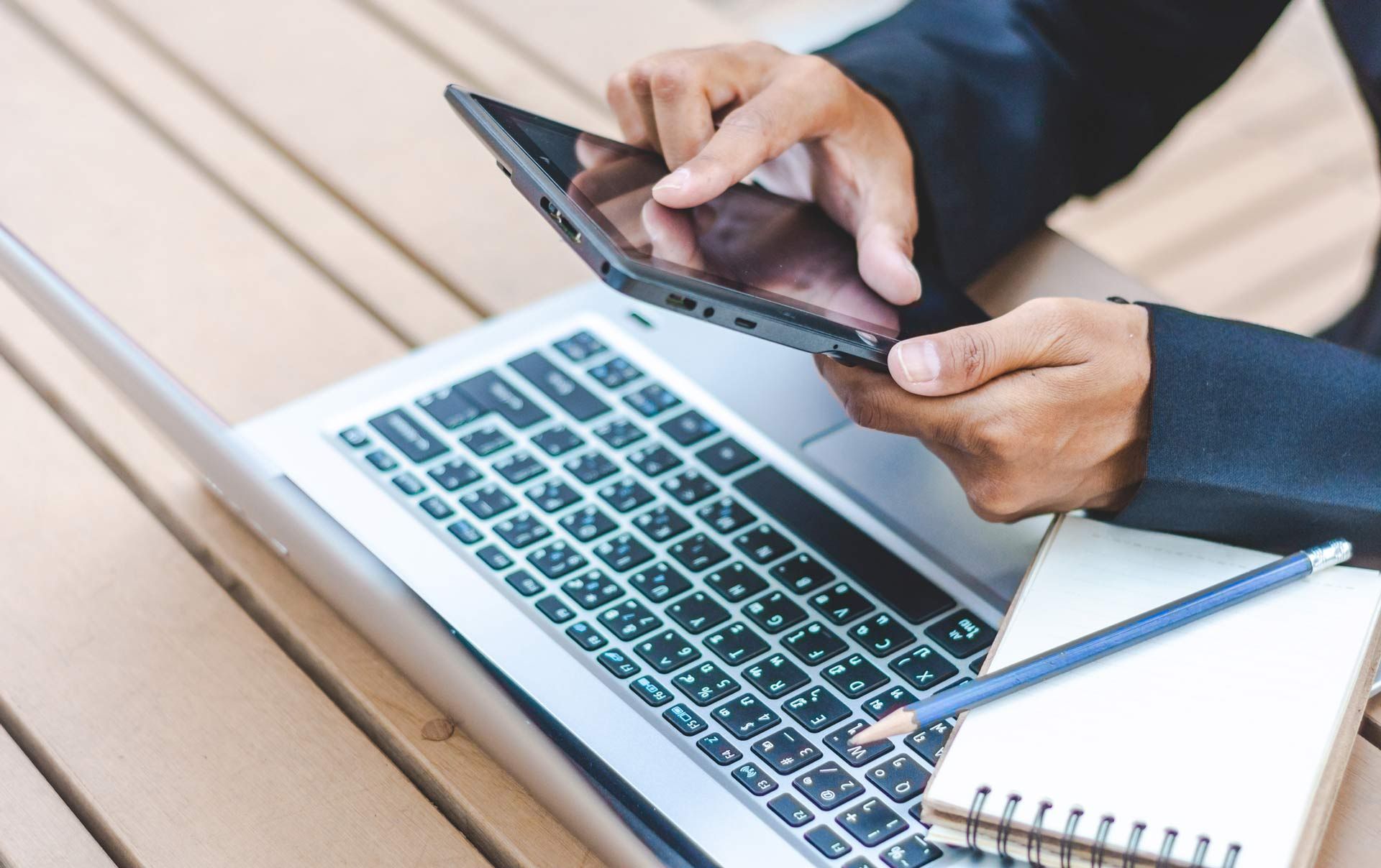 A person is using a tablet and a laptop on a wooden table.