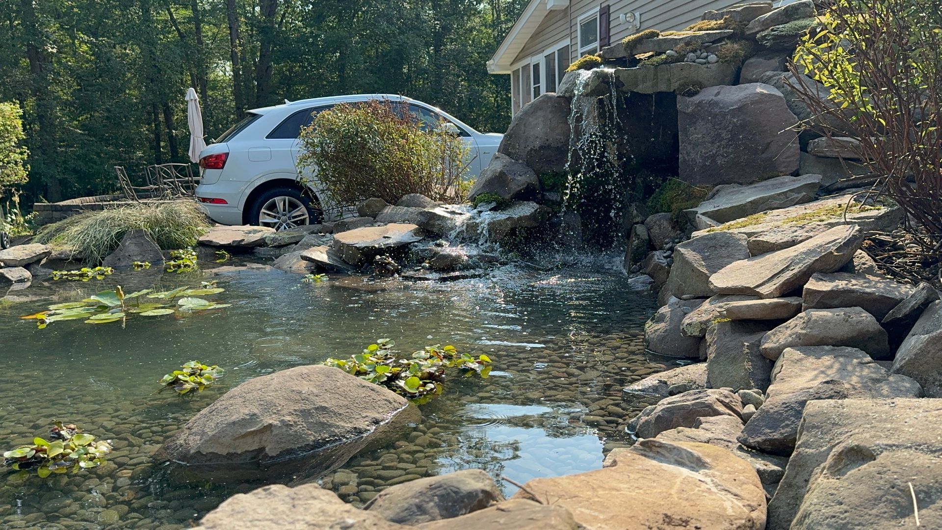 A car is parked in front of a house next to a pond with a waterfall.