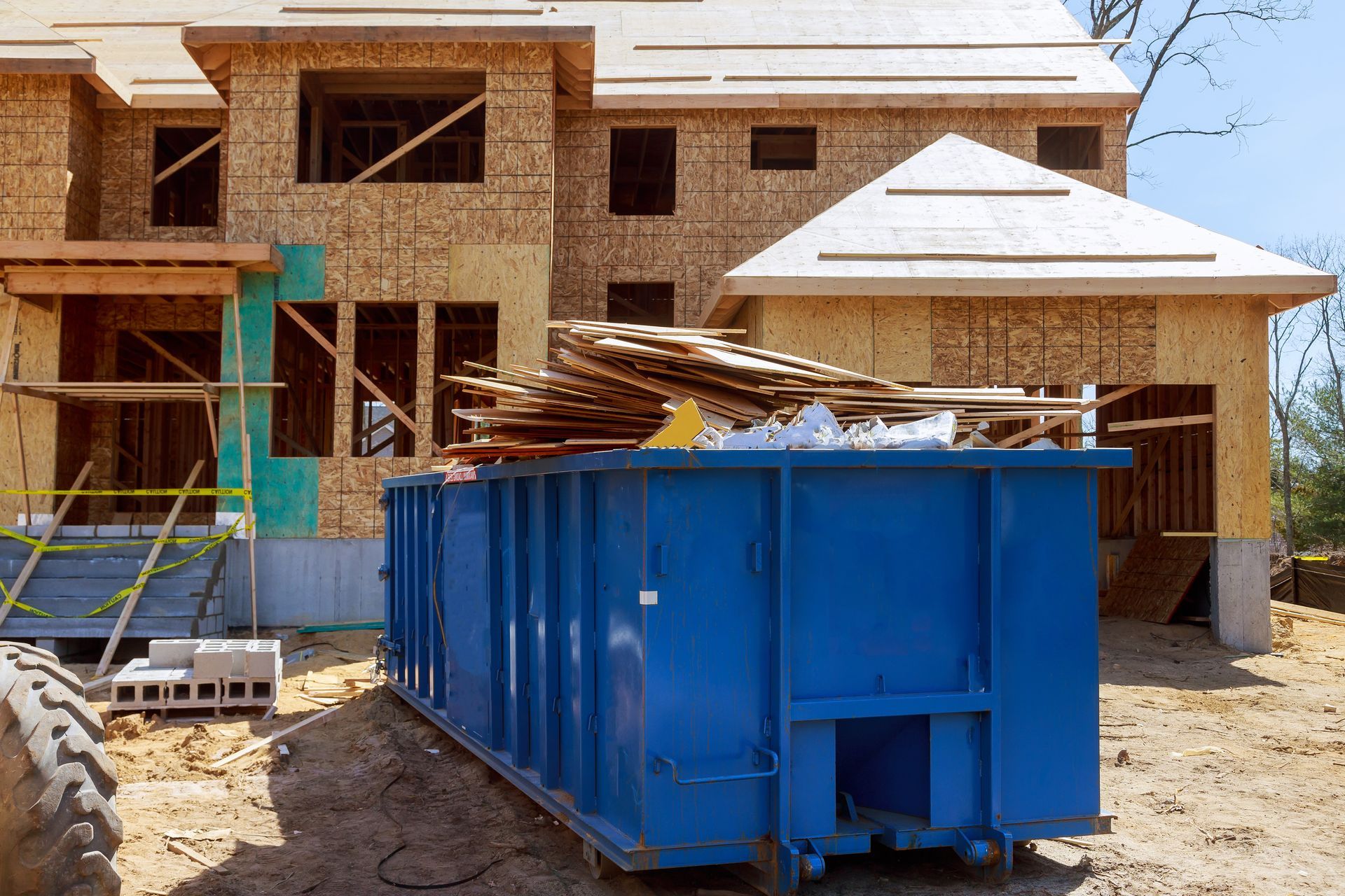 Blue construction dumpster in front of a partially built house, filled with wood scraps.