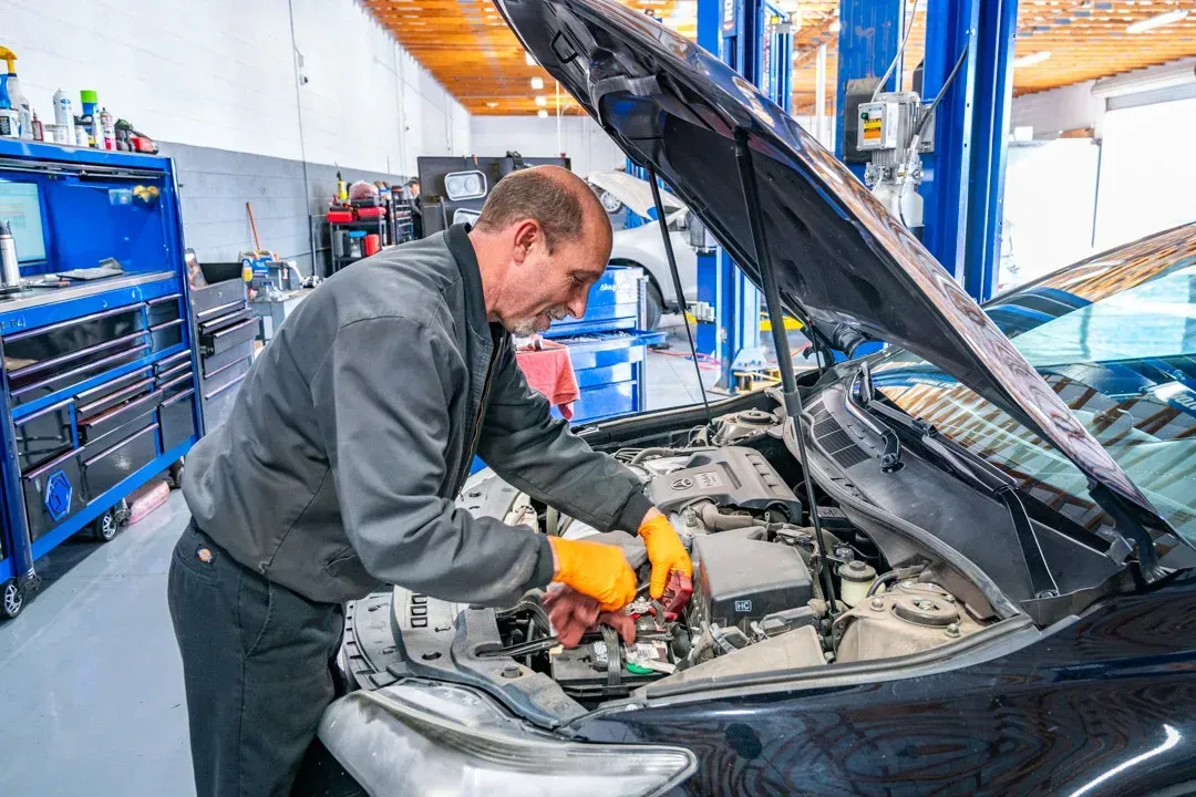 Mechanic working on a car engine in a garage; uses tools, wears gloves.