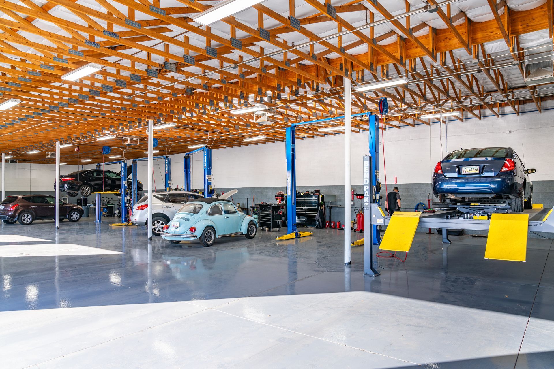 Car repair shop interior with vehicles on lifts, bright lighting, wood beam ceiling, and a shiny gray floor.
