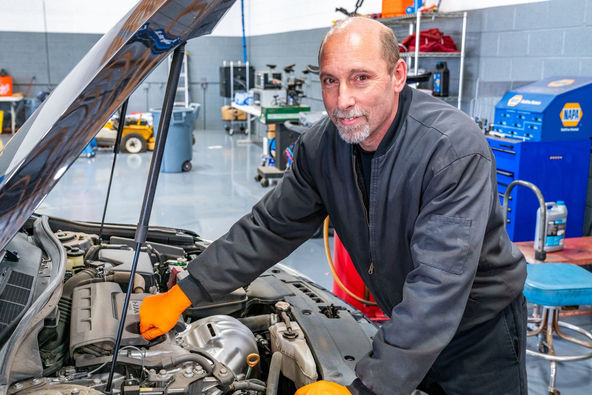 Mechanic in a garage, inspecting a car engine. Wearing orange gloves, he looks at the camera.
