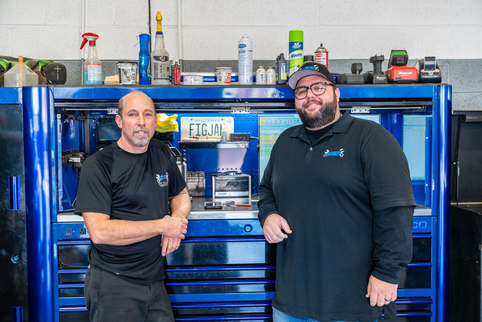 Two men stand by a blue tool chest in a shop. One wears a black shirt, the other a dark blue polo.