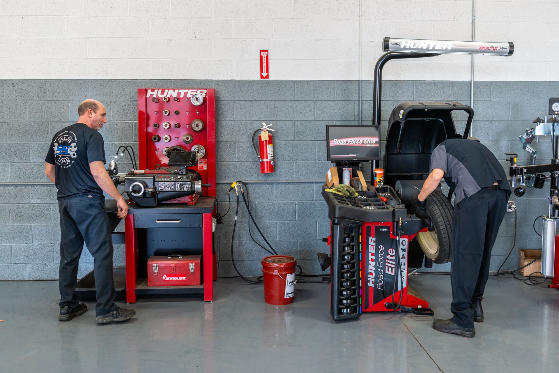 Two mechanics in a garage working on tires, with tire balancing and mounting equipment.