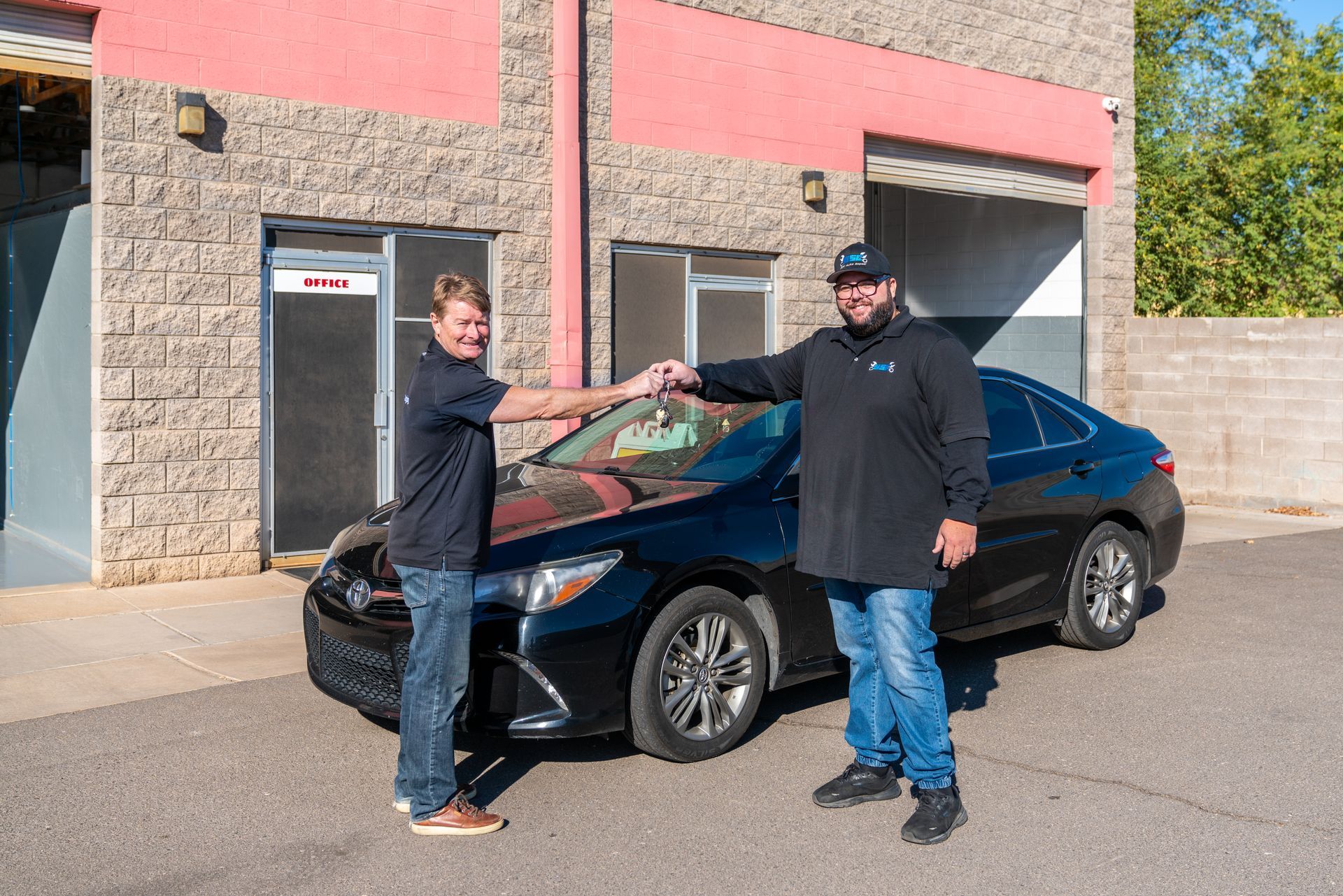 Two men fist-bump near a black car in front of a brick building with a garage door.