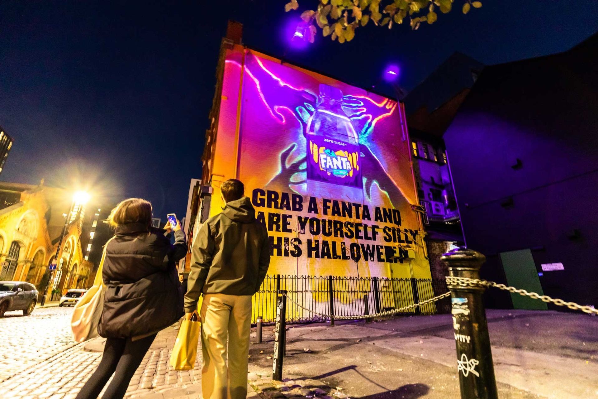 A couple of people are standing in front of a large mural on the side of a building. There is a mural on the side of a building that is illuminated by Absolute Events with UV light