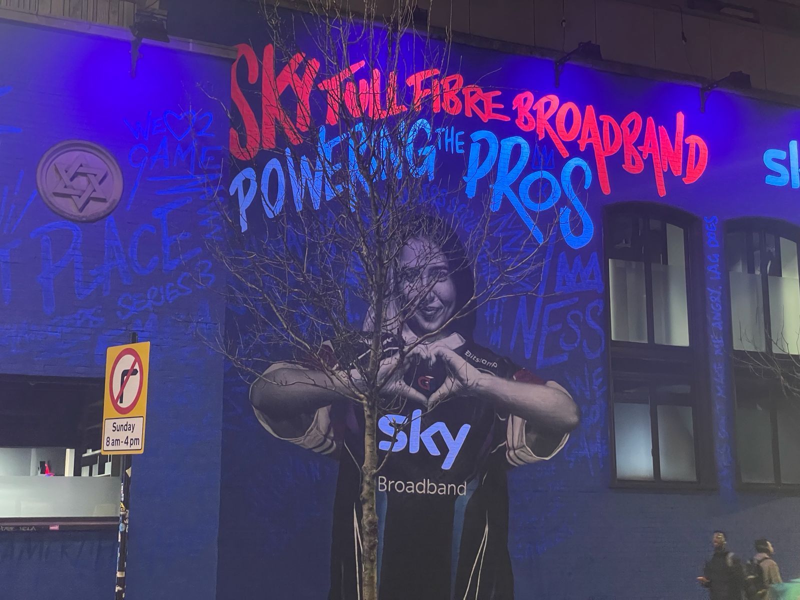 A blue building with a painting of a woman making a heart with her hands and the words sky on it, that is illuminated by Absolute Events with UV light