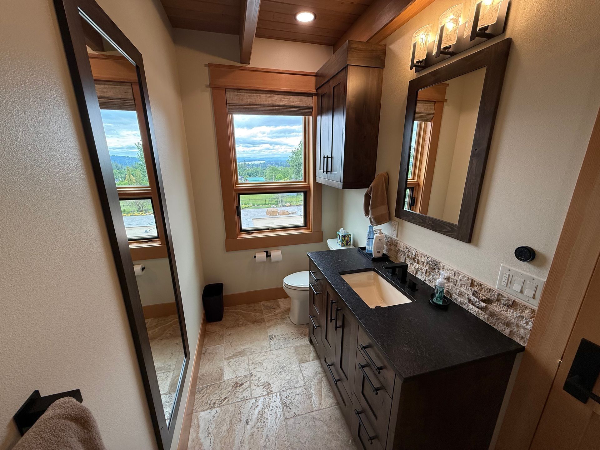 Custom bathroom vanity with black countertop, wood cabinetry, tile backsplash, and view window.
  