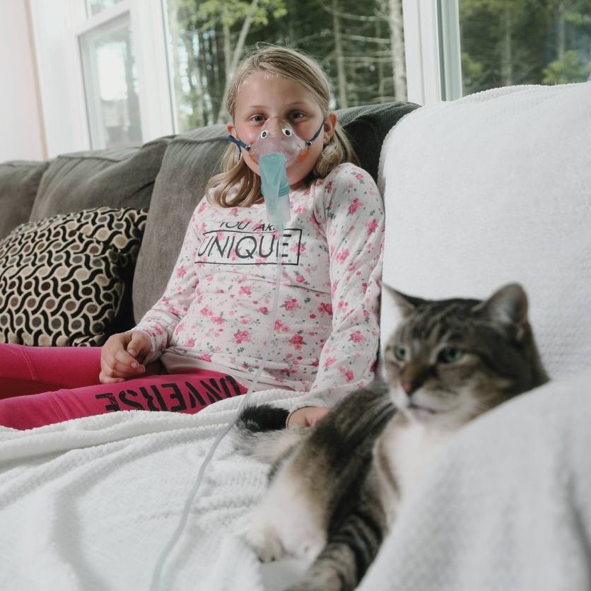 A young girl with CF sits on her couch with her cat as she received CF treatment