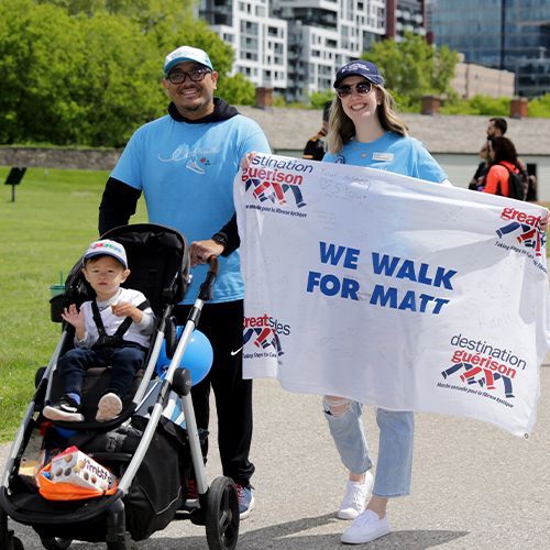 A baby rides in his stroller alonside his mom and dad who are holding a sign that says, We Walk for Matt