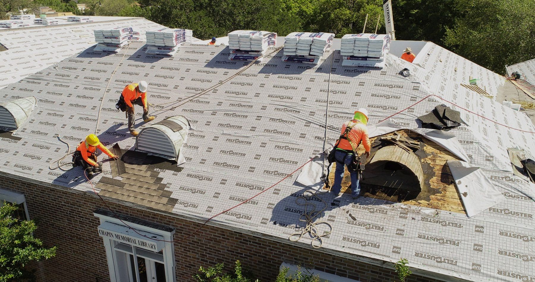 Roofers in orange vests working on a roof, surrounded by supplies, on a sunny day.