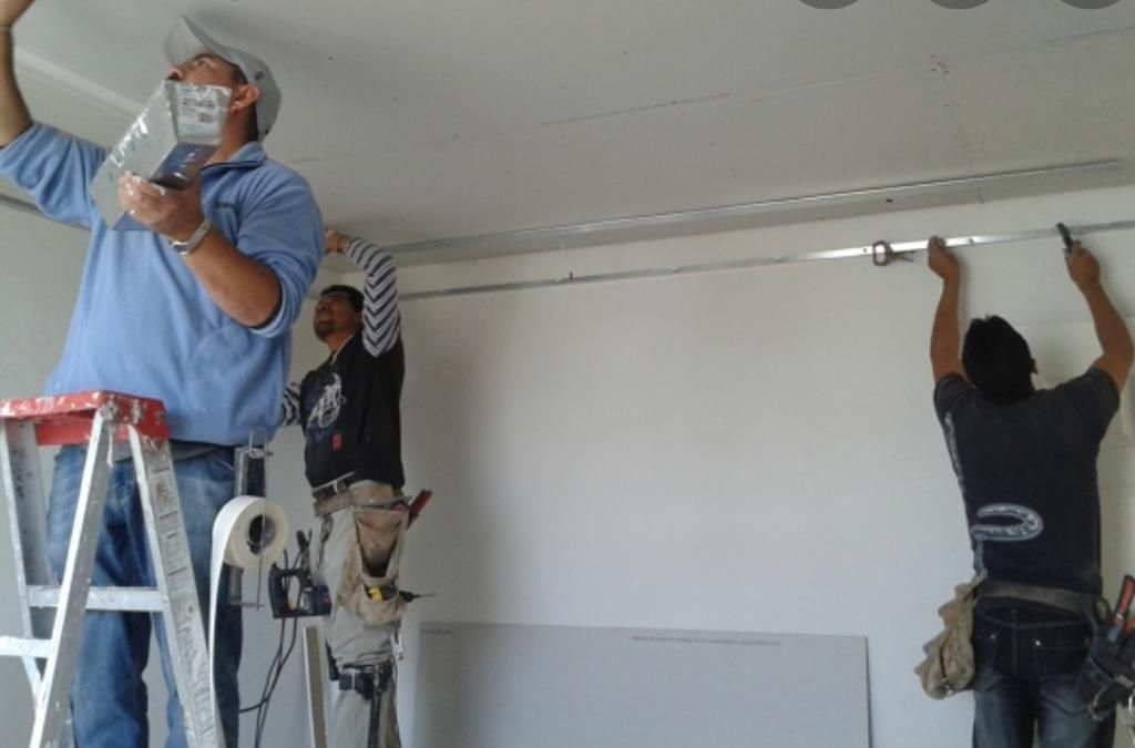 Three workers installing drywall on a ceiling. One on a ladder, two on the floor with tools. White walls.