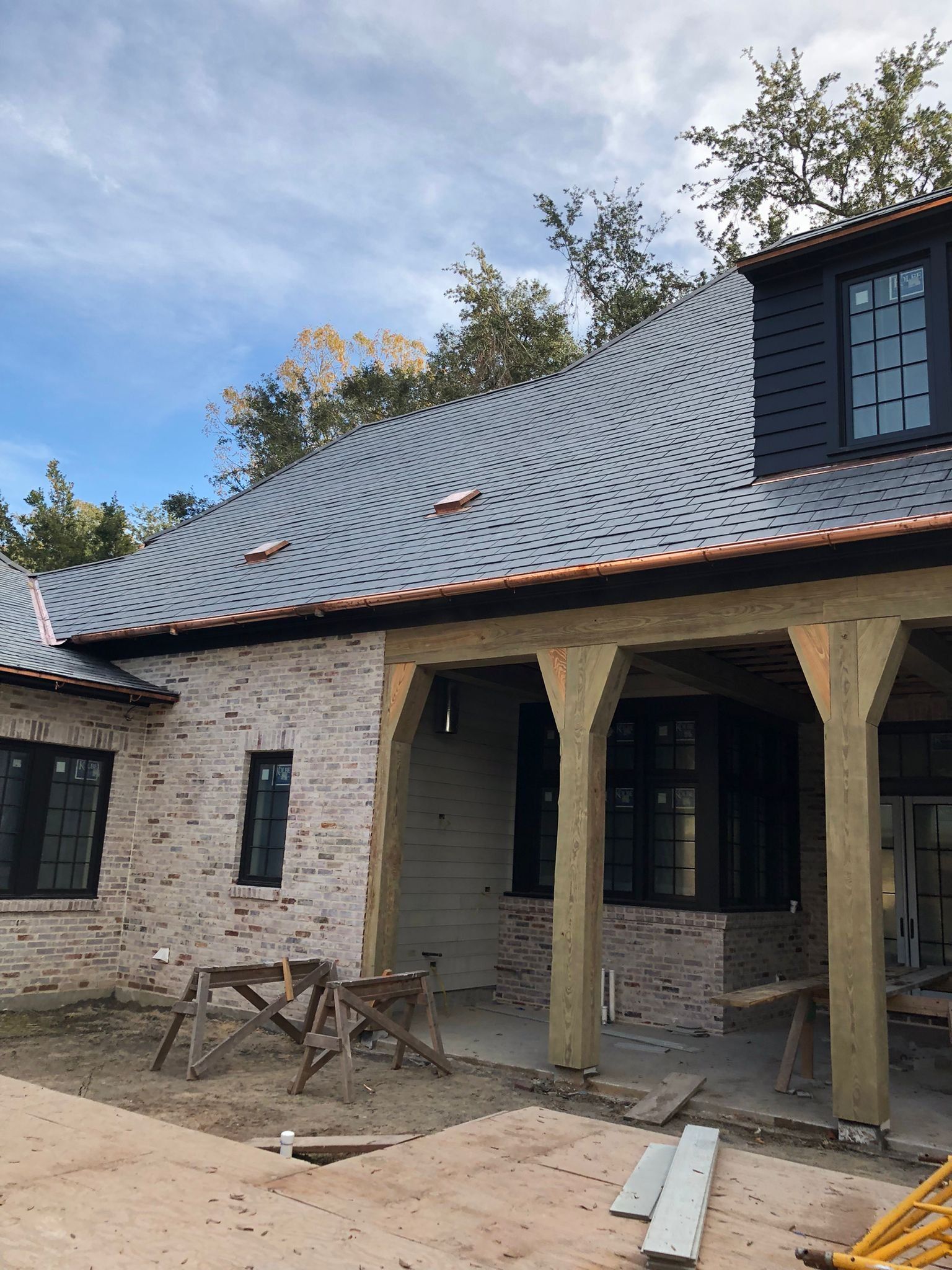 Brick house under construction with a wavy gray roof, dark trim, and wooden beams.