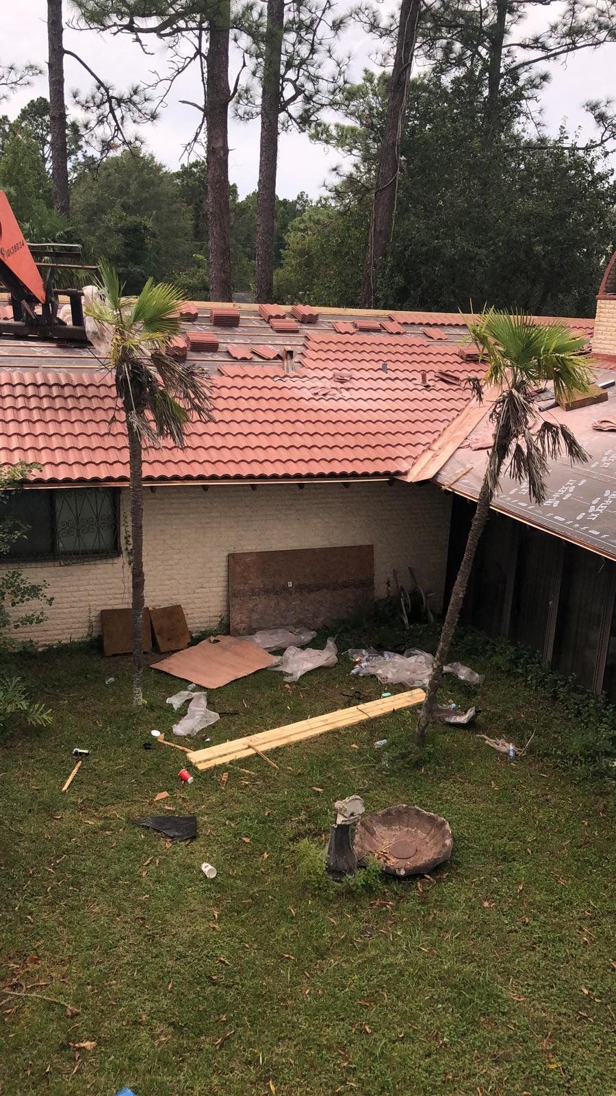 Damaged red-tiled roof on a house, with debris and missing tiles. Palm trees and backyard view.