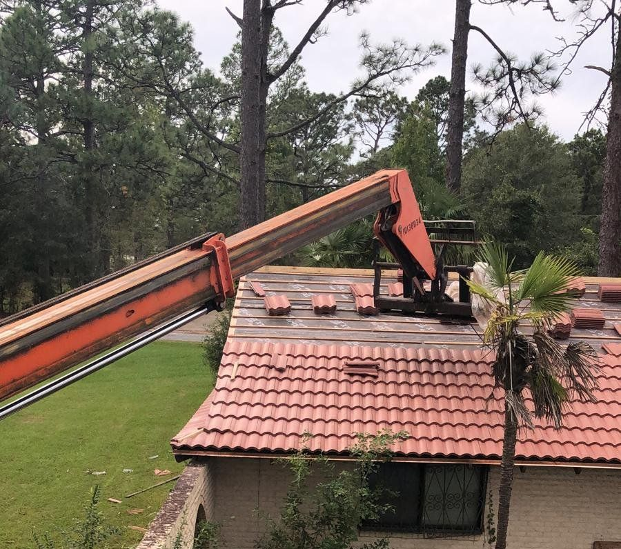 An orange lift removing roof tiles from a house with a red tile roof surrounded by trees.