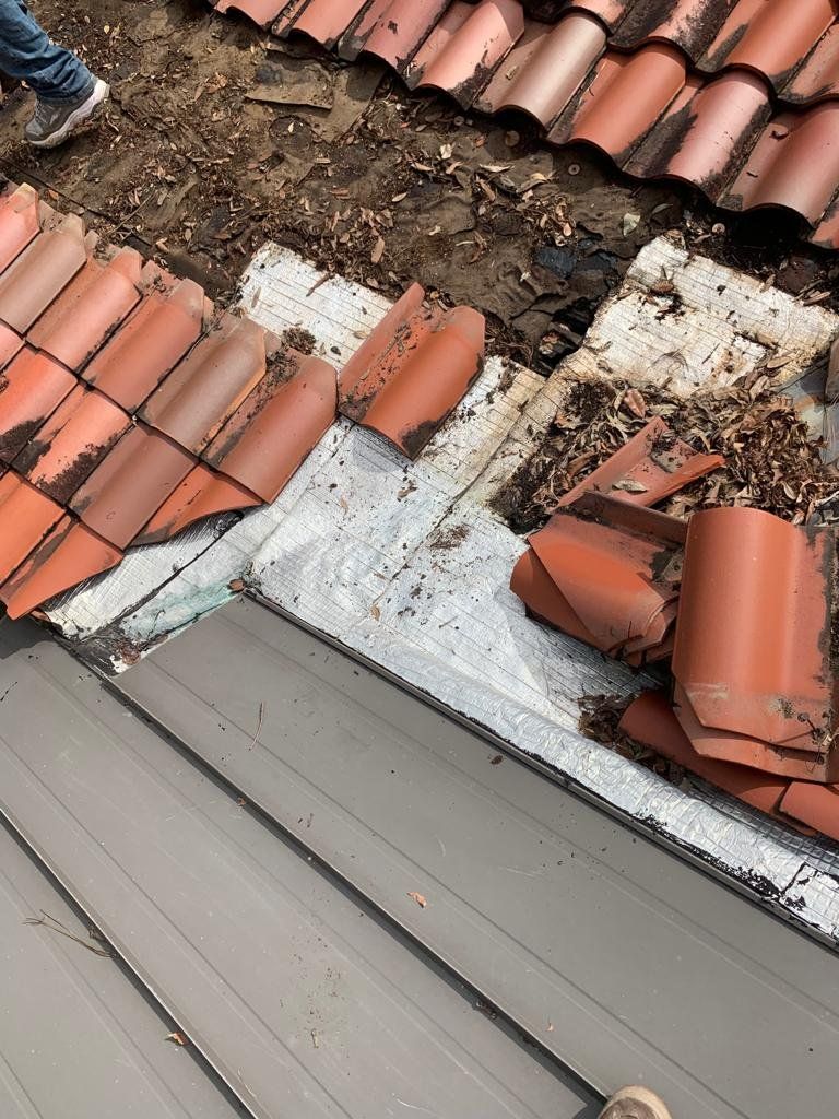 Roof tiles being removed, showing underlayment. Orange tiles, gray roofing, debris.