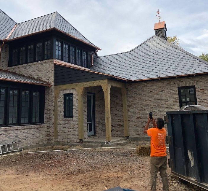 Man in orange shirt photographing a brick home with slate roof, copper gutters, and black windows.