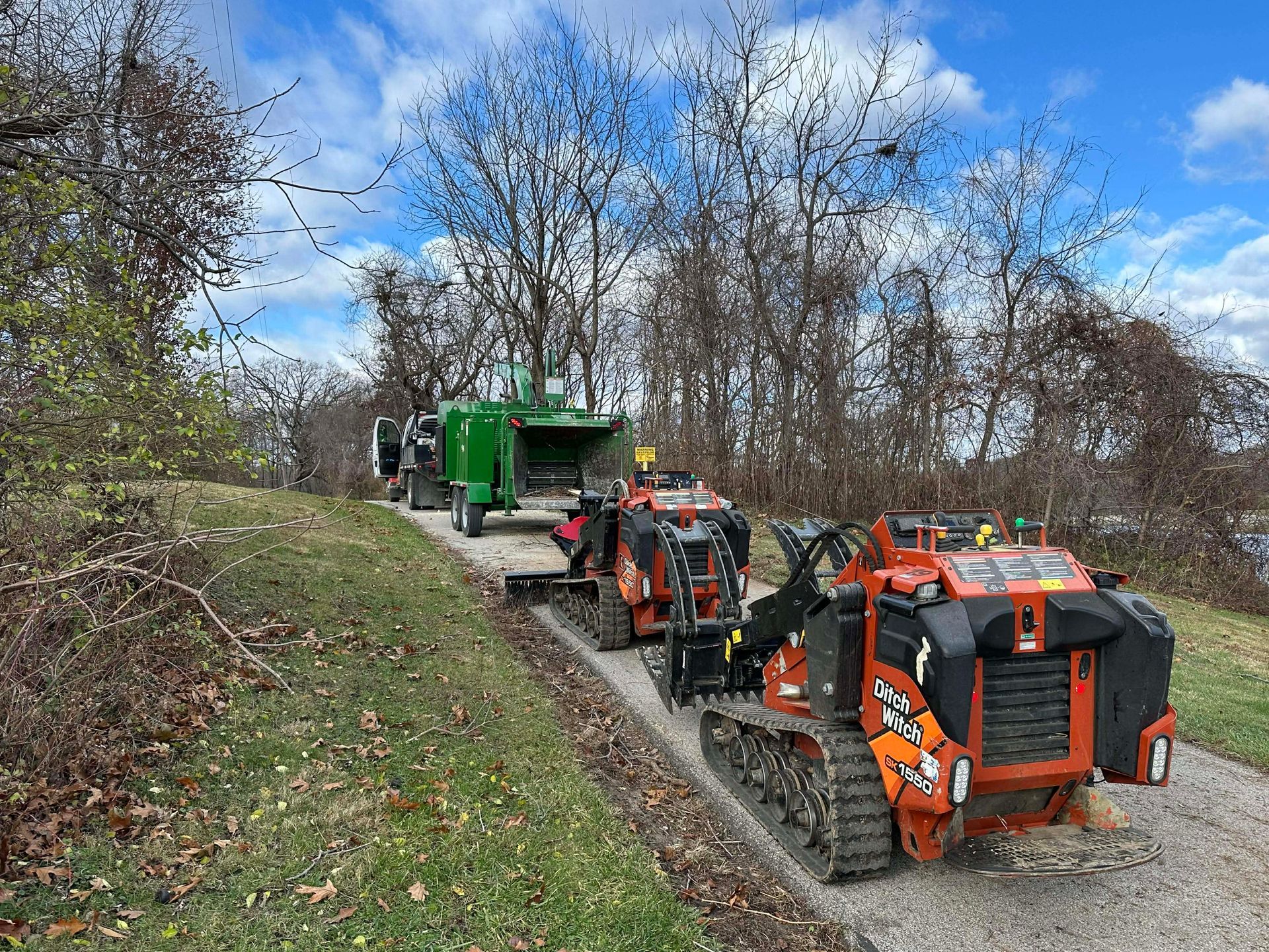 Two orange construction vehicles and a green machine on a gravel path near trees and water.