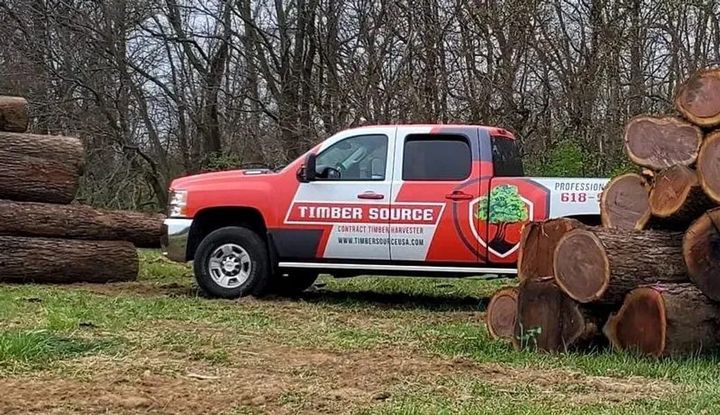 Red and white pickup truck with company logo parked near stacks of logs.