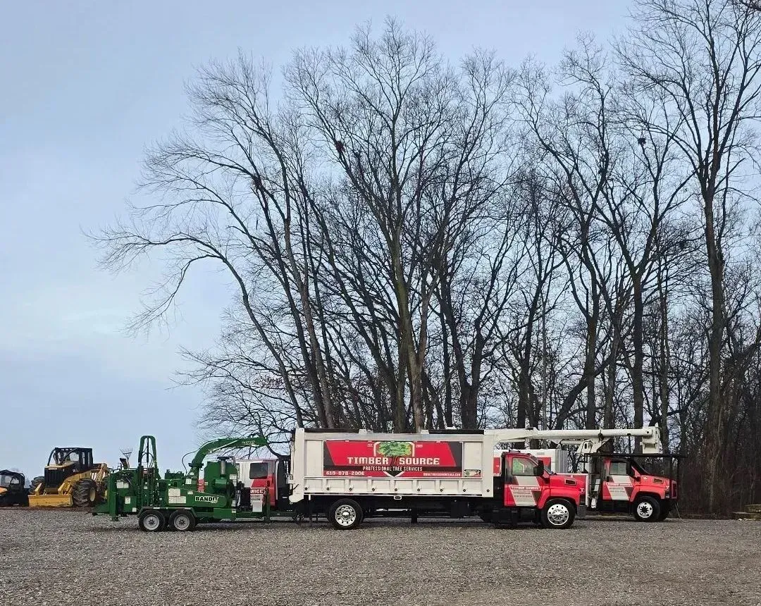 Truck and wood chipper in front of bare trees. White and red trucks with logo.