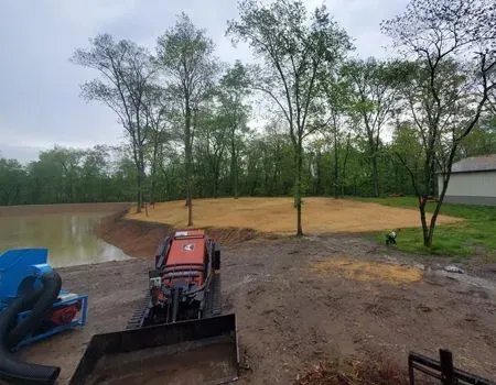 A small red tractor with a front-end loader on muddy ground near a pond and newly seeded earth, trees in the background.