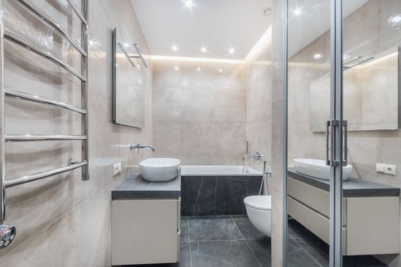 Modern bathroom with light beige tiles, a grey vanity with a vessel sink, a bathtub, and a wall-mounted towel warmer.
