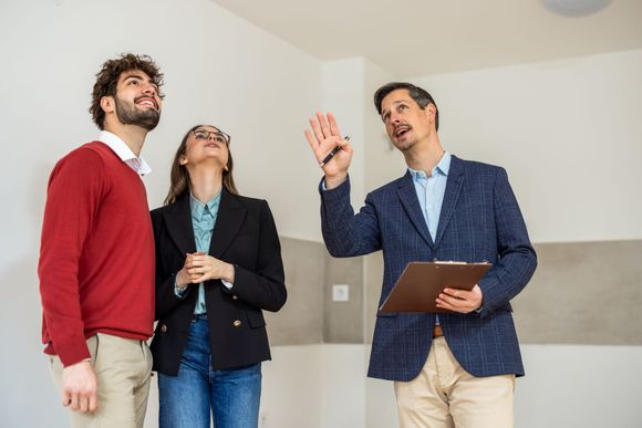 A real estate agent points toward the ceiling while explaining details to a couple inside an unfurnished room.