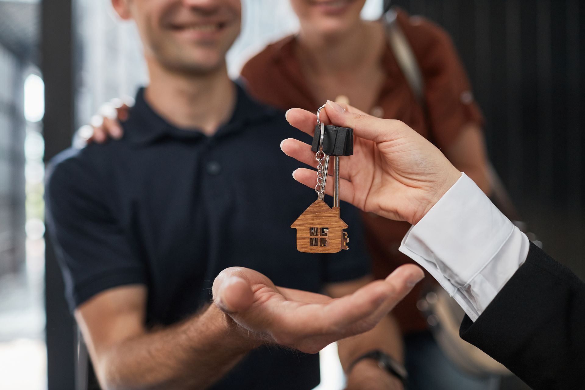 Person handing house keys to a couple; smiling, blurred background.