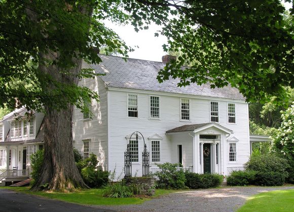 White two-story house with a porch and ornate entrance, shaded by a large tree, on a green lawn.