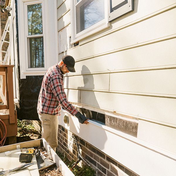 A person in a plaid shirt and cap is repairing the exterior wall of a house, inspecting the siding. A person in a plaid shirt and cap is repairing the exterior wall of a house, inspecting the siding.