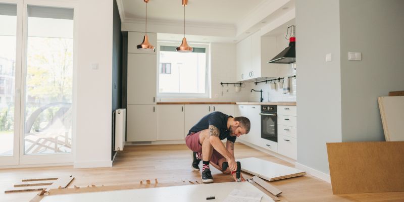 A person assembles furniture in a bright kitchen. They kneel on the floor with tools and white cabin