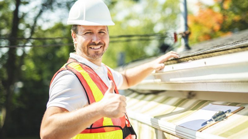 Man in hard hat and vest giving thumbs up while working on a gutter.