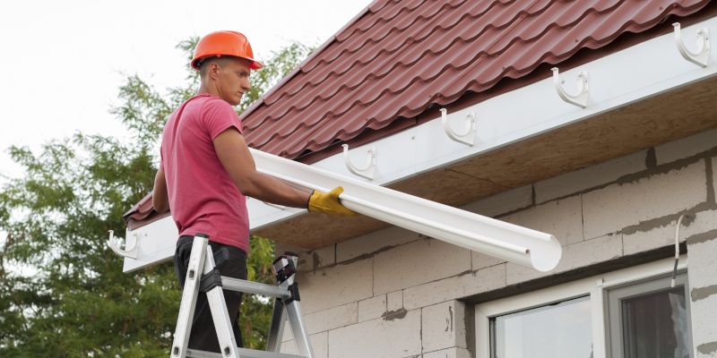Construction worker on a ladder installing a white gutter on a house roof.