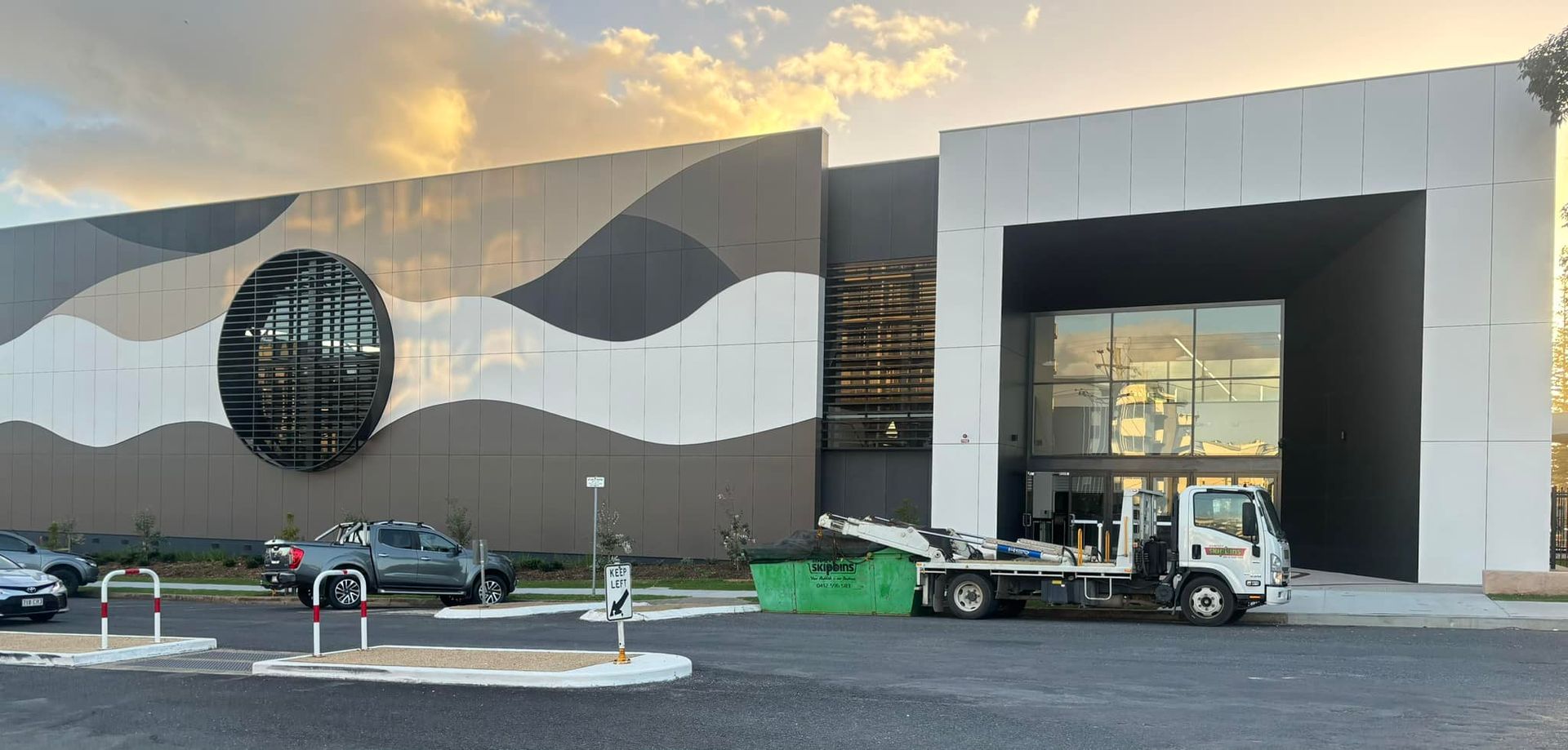 A White Flatbed Truck Transporting A Green Skip Bin Parked In Front Of A Building — Kempsey Skip Bins In Kundabung, NSW