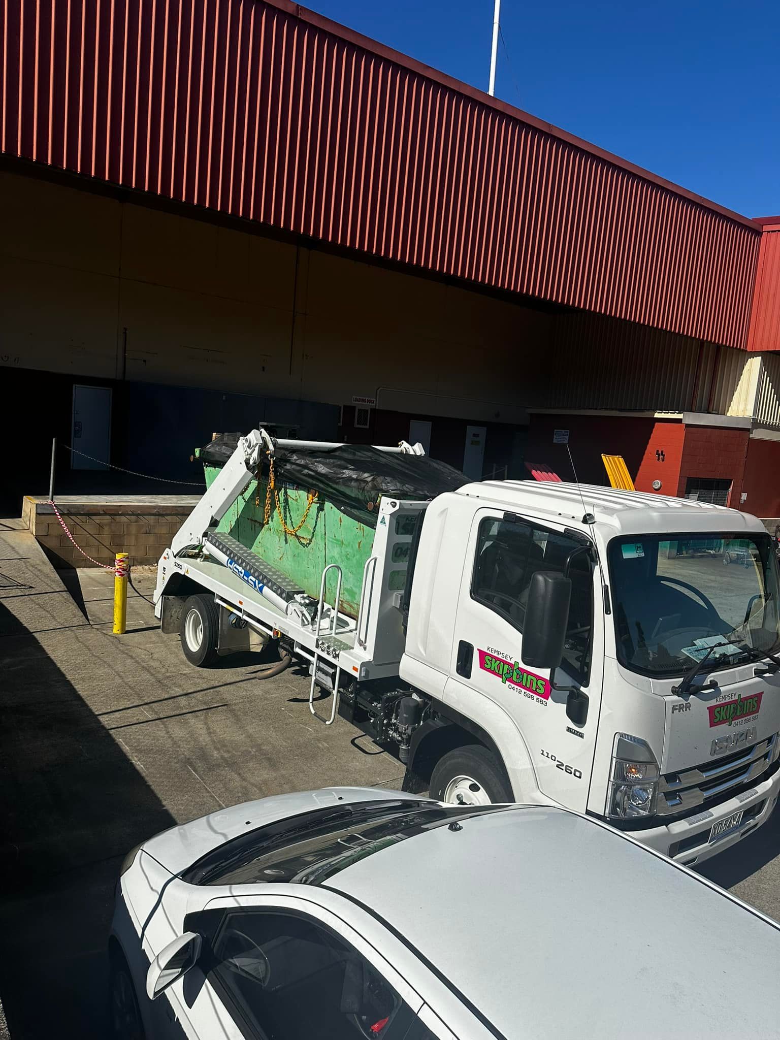 White Flatbed Truck Carrying A Green Skip Bin Full Of Rubbish — Kempsey Skip Bins In Smithtown, NSW