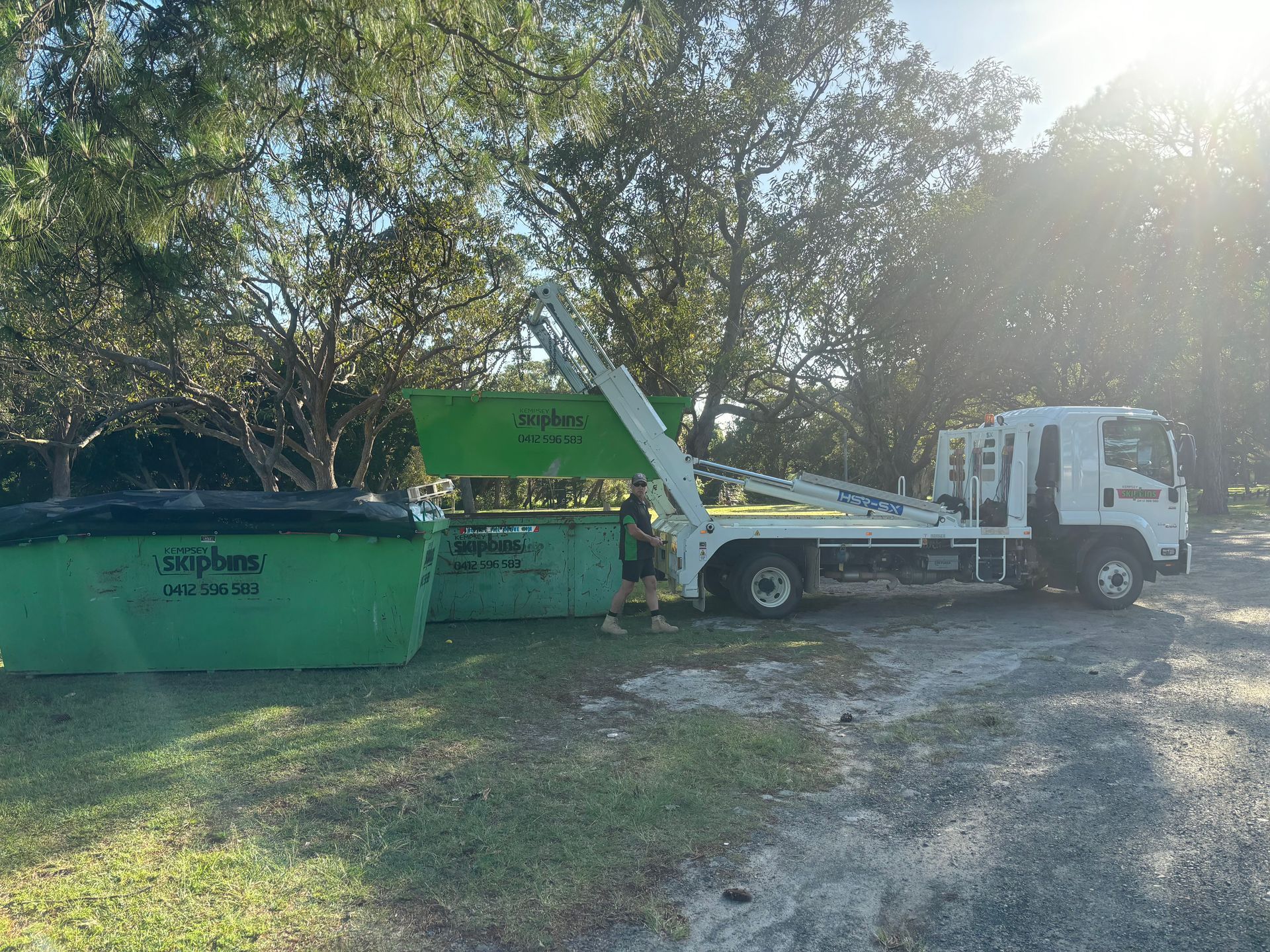 A Truck with Lifting Arms Loads a Green Dumpster on A Grassy Area Near Trees — Kempsey Skip Bins In Crescent Head, NSW