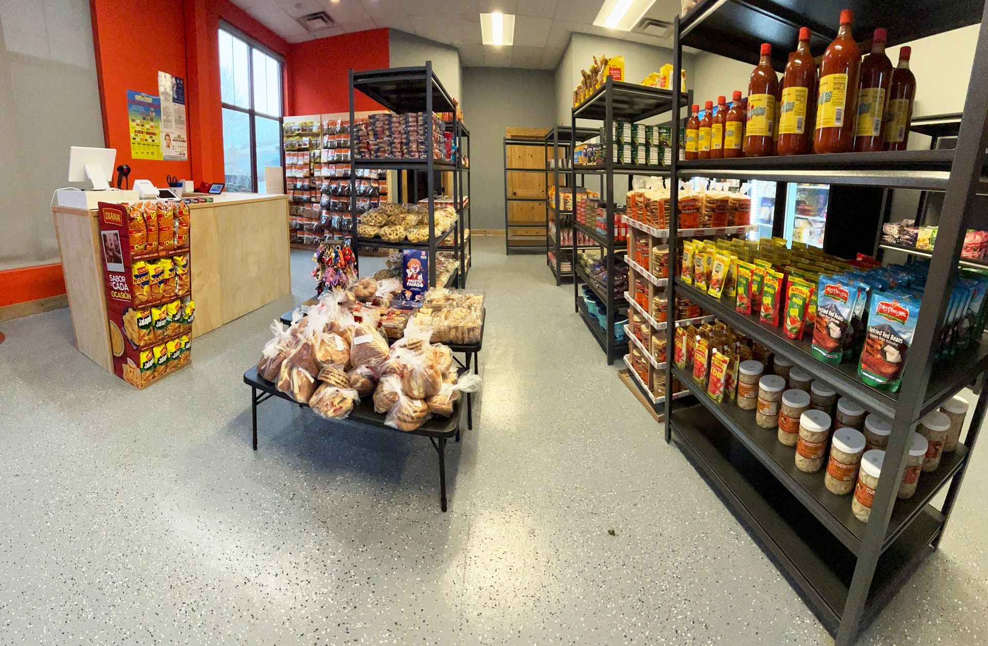 Interior of a small grocery store with shelves stocked with food items. Gray floor, orange walls, and a checkout counter.