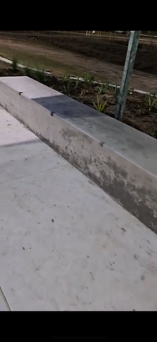Concrete bench in a park setting, with a paved walkway in the foreground and greenery in the background.