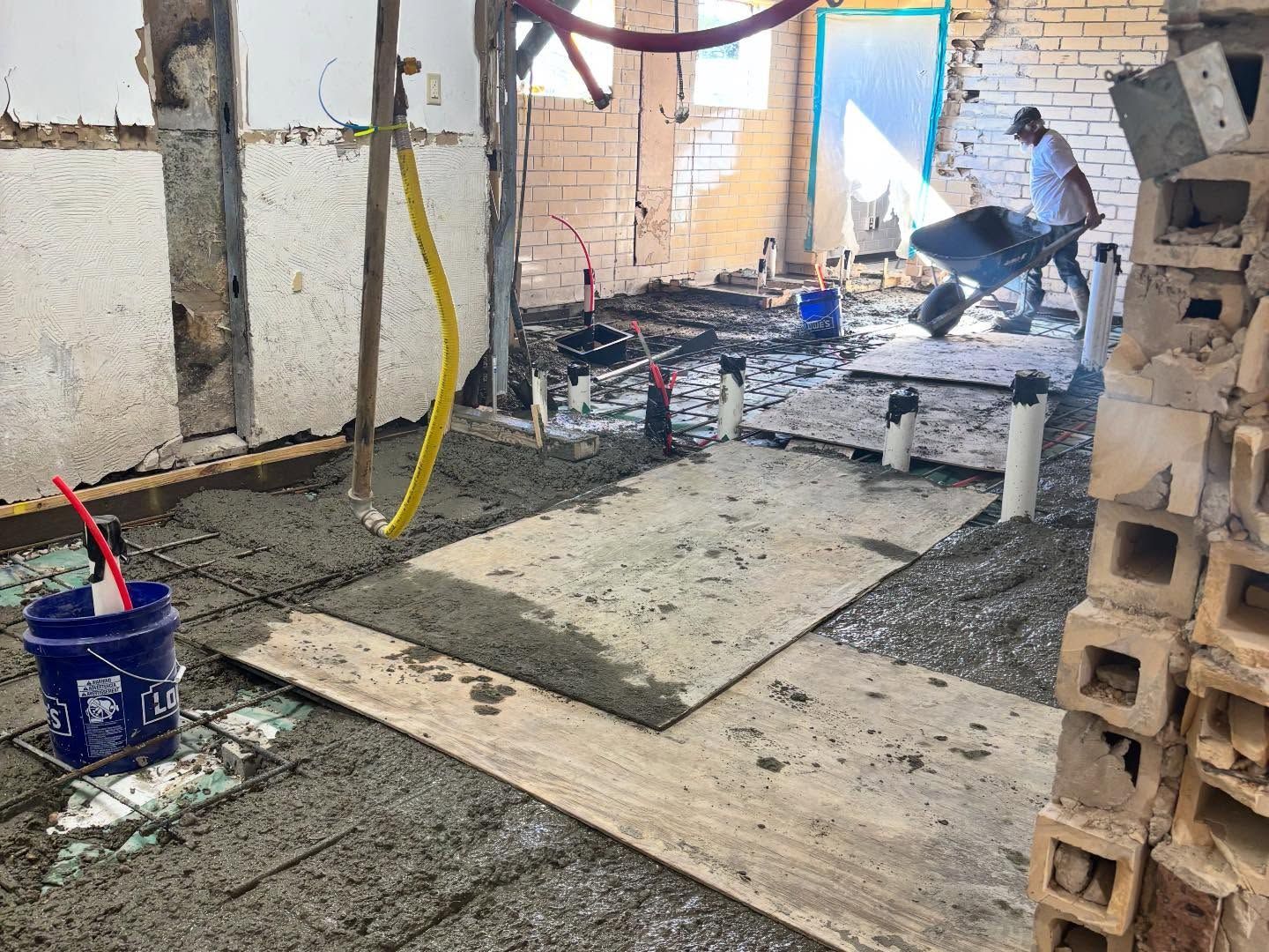 Construction worker pouring concrete; interior room with exposed pipes, white brick walls, a blue bucket, and a wheelbarrow.