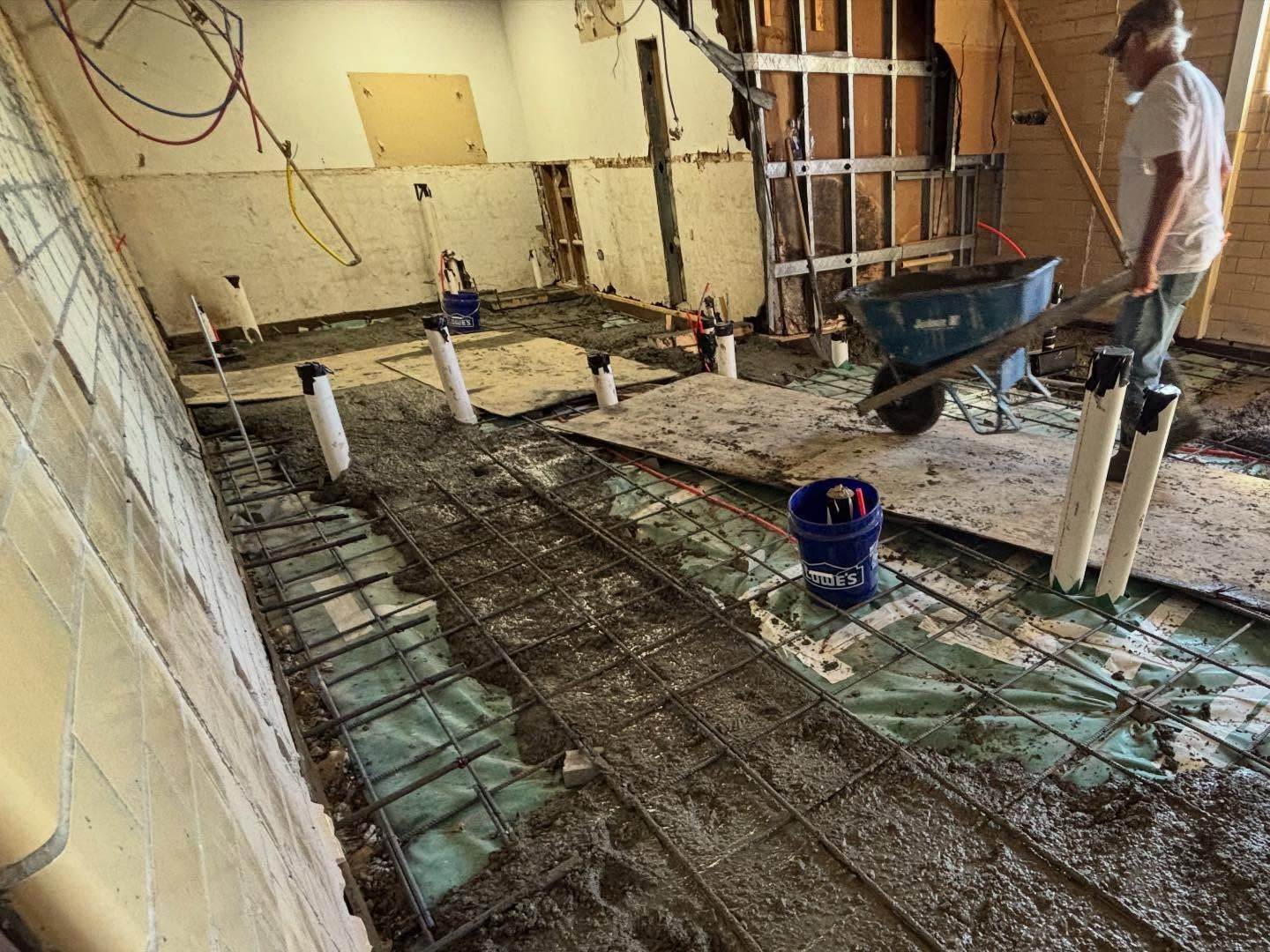 Construction site: man pushing wheelbarrow, rebar and plumbing pipes visible on floor, exposed wall.