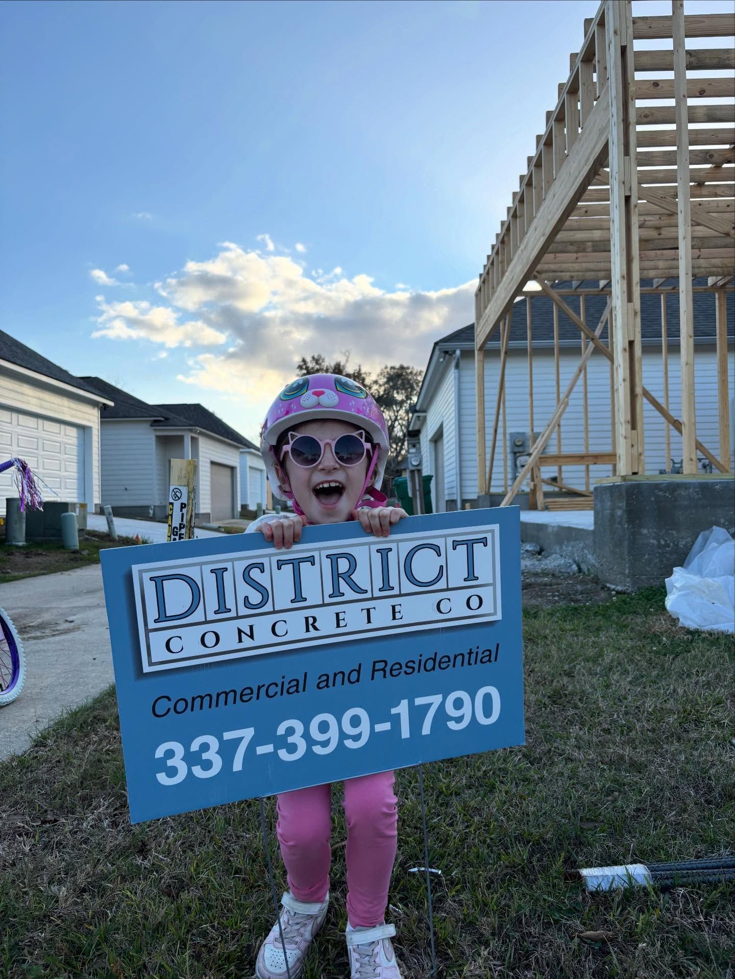 Child in pink outfit and helmet holding a District Concrete Co. sign, smiling in front of a building under construction.
