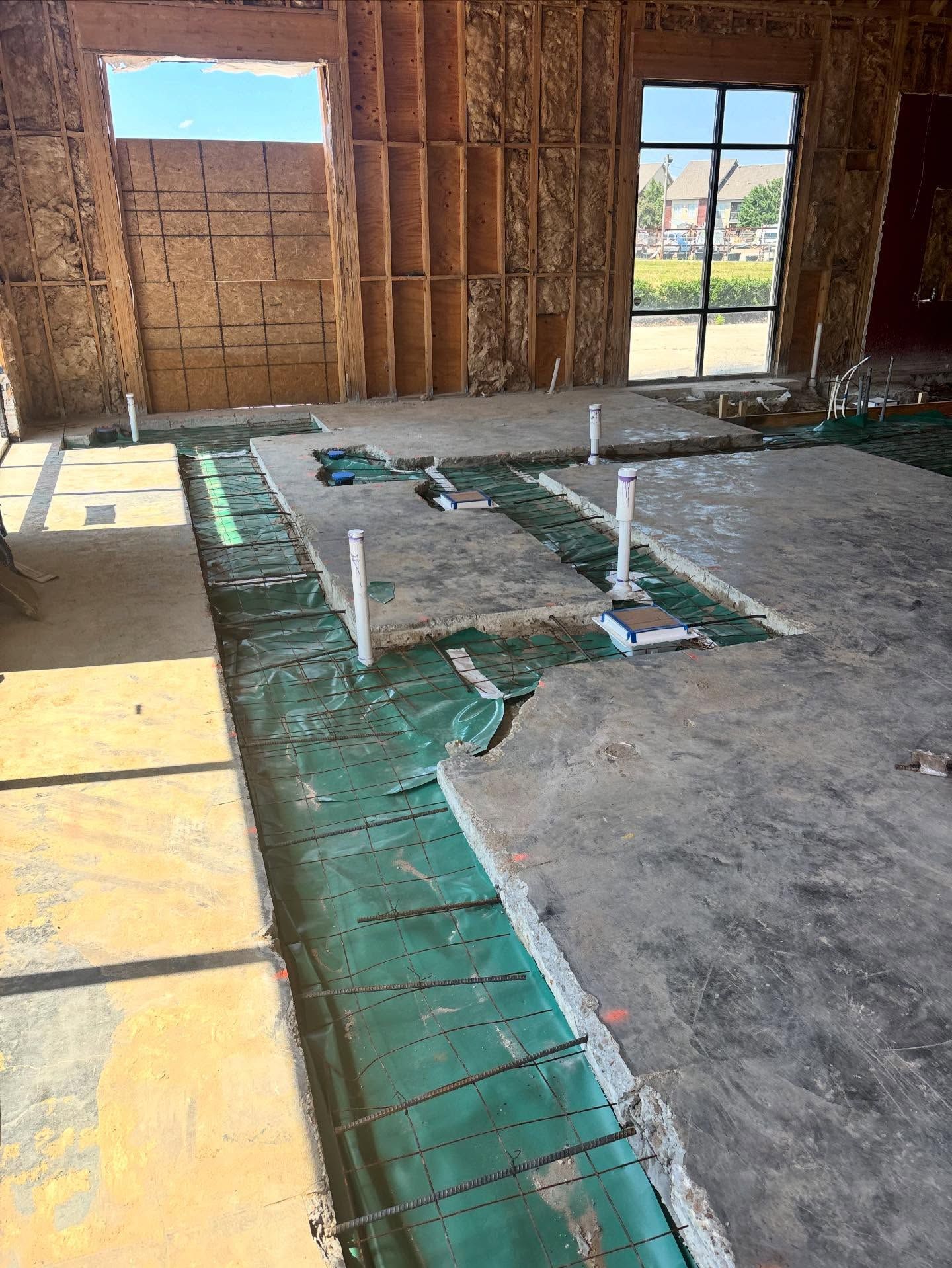 Interior construction of a building. Concrete floor with plumbing pipes and rebar exposed, wooden framework, large windows.