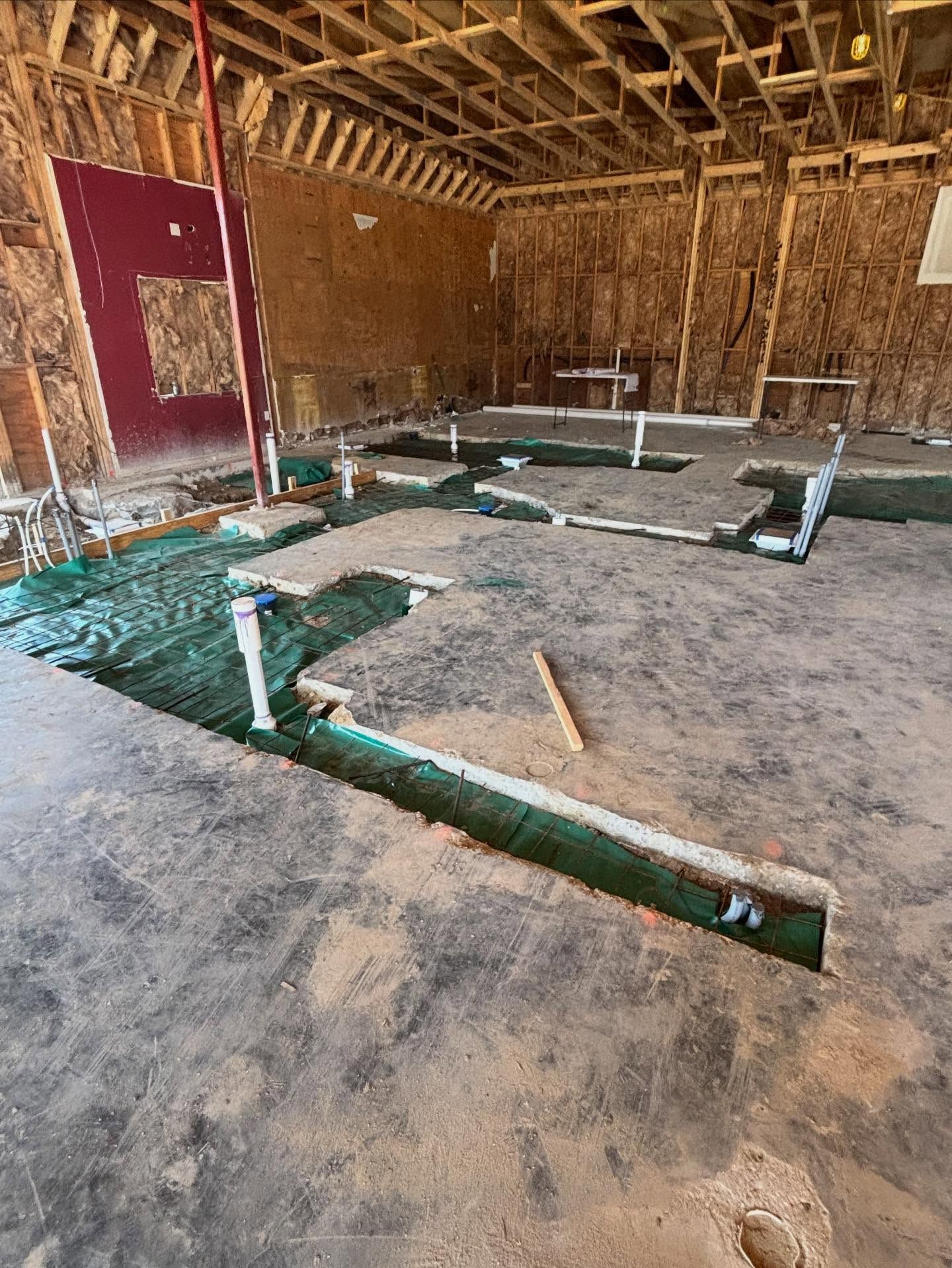 Construction site interior with plumbing pipes in trenches. Green mesh laid down. Unfinished walls.