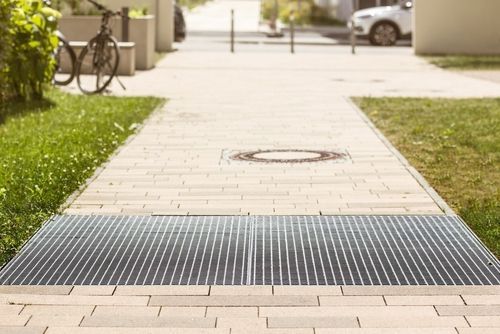 Grating on a paved walkway, with grass and buildings on either side, bicycle and car in the background.
