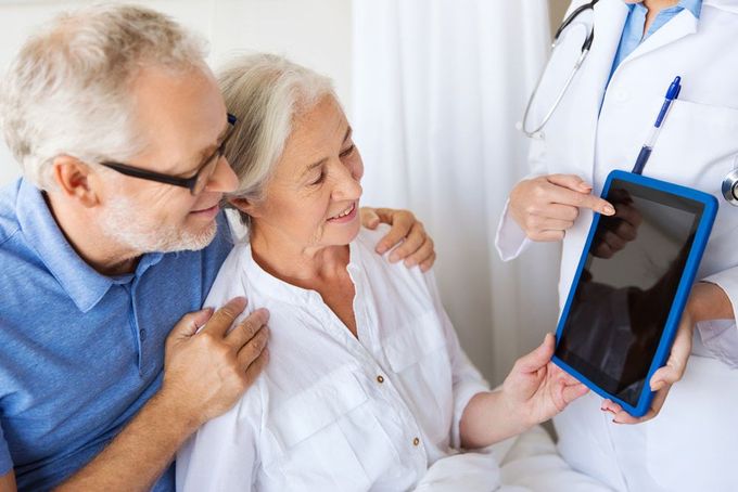 An Elderly Couple Is Sitting In A Hospital Bed While A Doctor Shows Them A Tablet — Miami Denture Clinic In Burleigh Heads, QLD