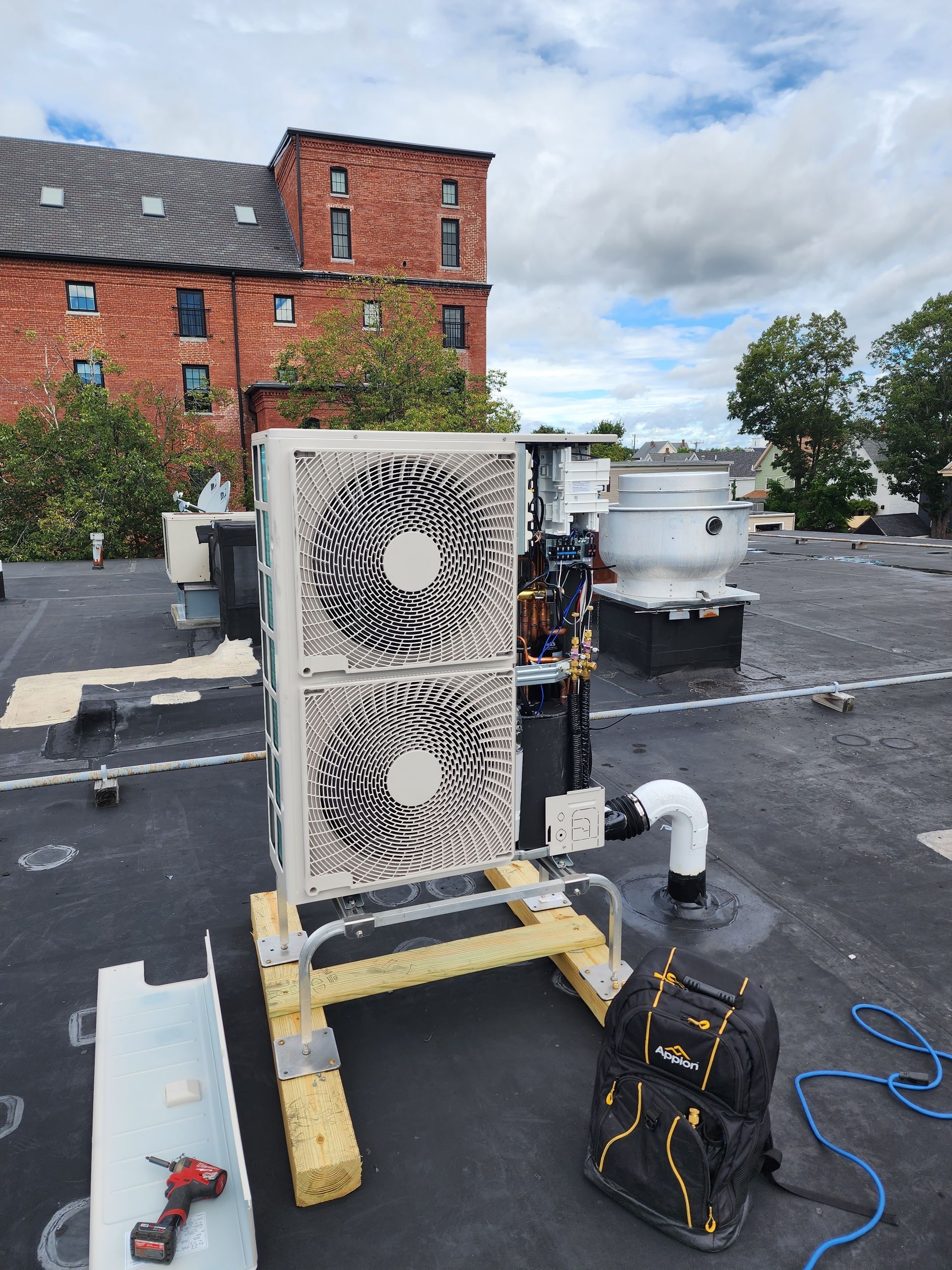 HVAC unit on a flat roof, supported by wooden frame, with brick building background and cloudy sky.