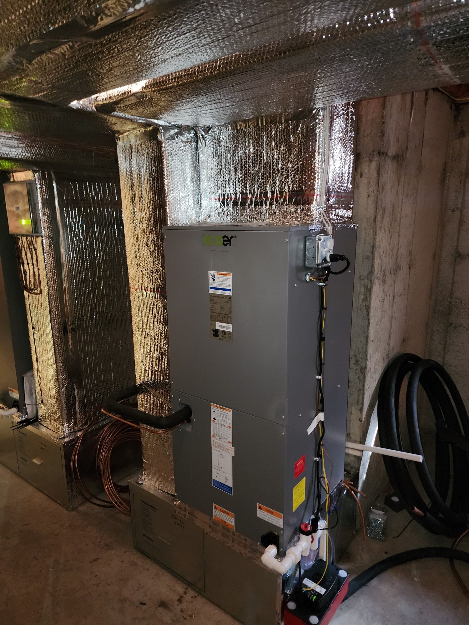 HVAC unit in a basement, ductwork is wrapped in silver insulation, next to a concrete wall.