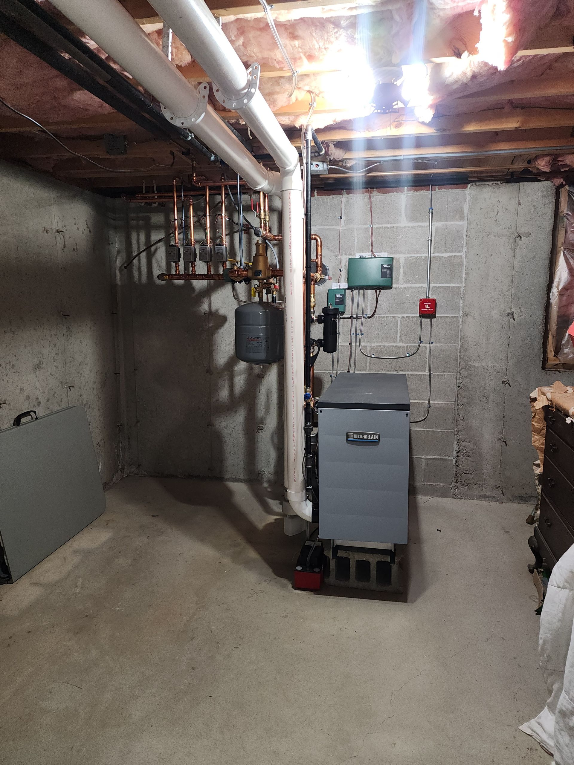 Basement with a grey boiler, pipes, and a fire alarm on a concrete wall.