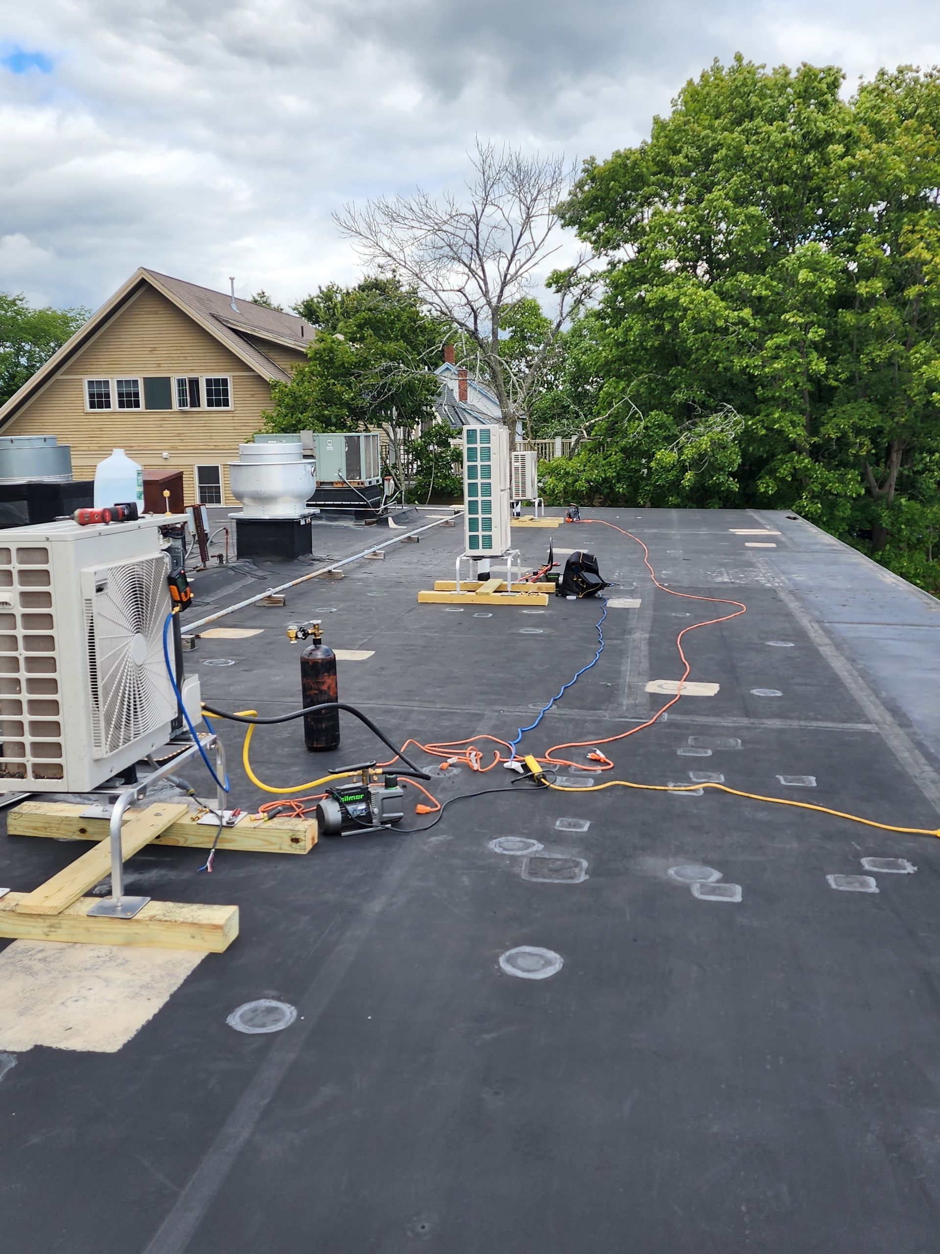 Rooftop with HVAC units, wires, and equipment. A house and trees are in the background under a cloudy sky.