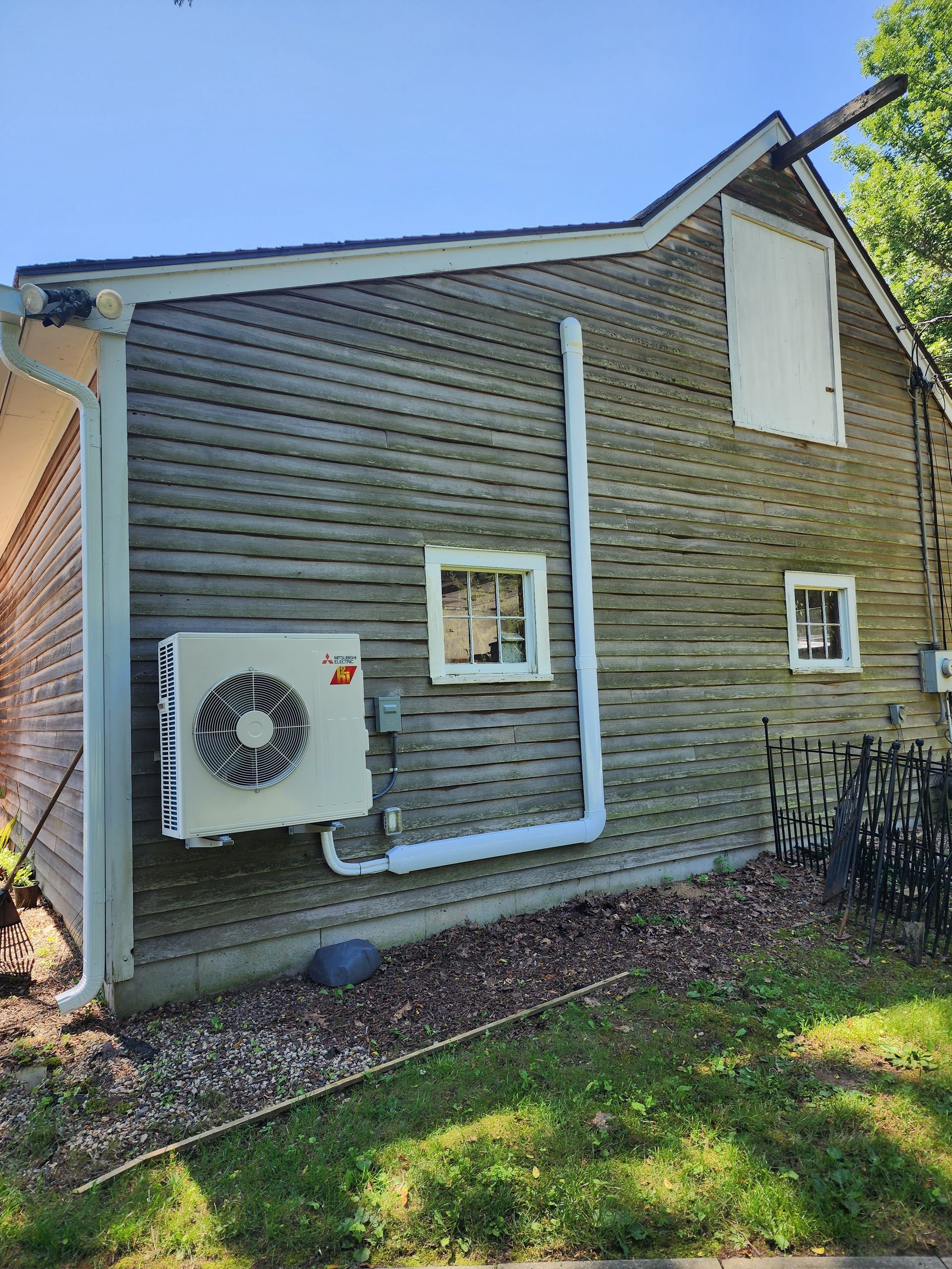 Exterior wall of building with a heat pump unit. White siding with a window, a door, and white piping.